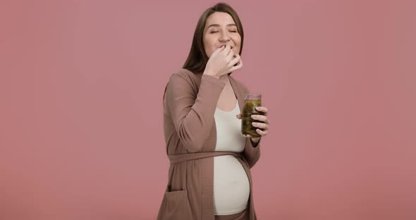 Studio Portrait of Young Pregnant Lady Enjoying Pickles Eating Them From Jar Pink Background