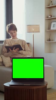 A Laptop with a Green Screen Stands on a Table Against of a Woman Reading a Book