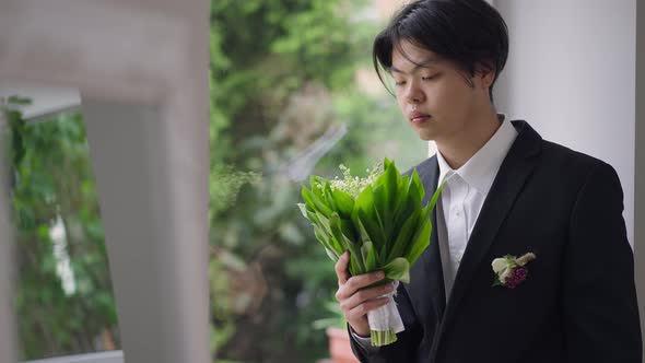 Romantic Confident Asian Groom Admiring Bouquet of Lilies of the Valley and Looking Out the Window