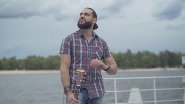 Portrait of Handsome Caucasian Musician Standing with Guitar on Urban Embankment and Looking Around