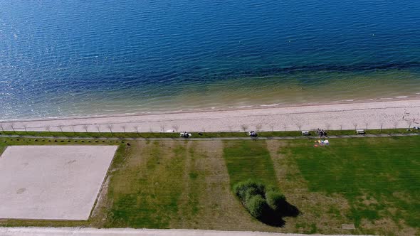 Lake beach with people sunbathing and bathing, garden and walking area. Overhead drone shot travelin