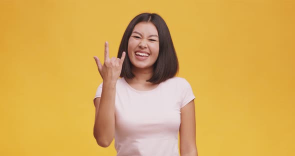 Carefree Asian Woman Demonstrating Rock and Roll Hand Gesture, Punk Sign, Orange Studio Background