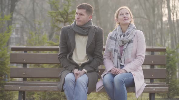 Portrait of Shy Adult Caucasian Couple with Age Difference Sitting on Bench in City Park. Younger