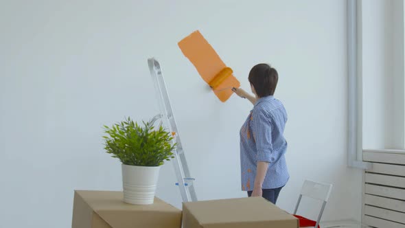 Flat Renovation Concept. Happy Middle-aged Woman Painting White Wall with Paint Roller, Orange Paint