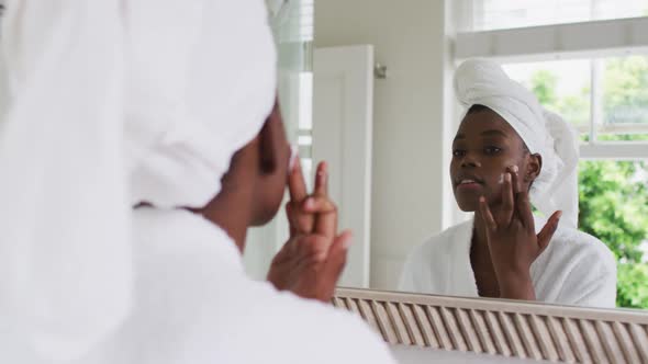 African american woman in bathrobe applying face cream while looking in the mirror at home