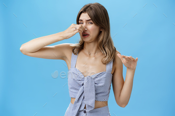 oung slim woman with tattoo squeezing nose with two fingers not sniff disfusting reek crying from stink standing intense over blue background.