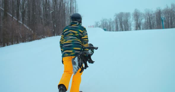 Snowboarder Climbs the Mountain in a Ski Suit and with a Snowboard