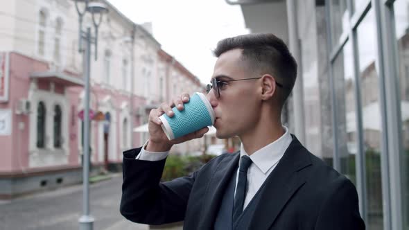 Elegant Man in Suit with Dark Sunglasses Drinks a Cup of Coffee and Looks Around