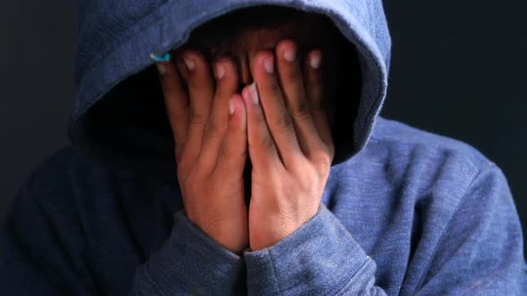 Sad Man in Hood Cover Face with Hands Isolated in Black.