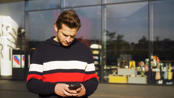 An adult men wearing a coloured hoodie is using his smartphone in front of a shop in Amsterdam, Neth