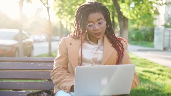 African American Woman Sitting on a Bench in City and Using Laptop