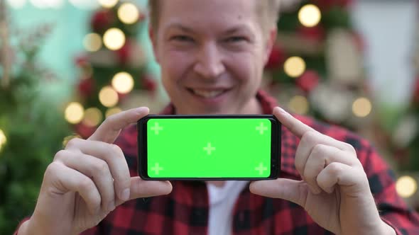 Young Happy Hipster Man Showing Phone Against Christmas Trees Outdoors