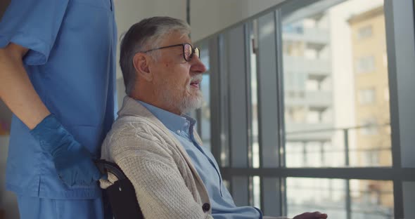 Senior Man in Wheelchair with Nurse Taking Care Looking Through Hospital Window