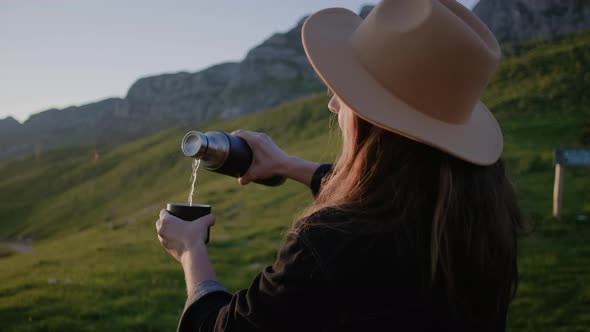 Back view of hipster woman in hat pours hot tea and drinking from thermo cup