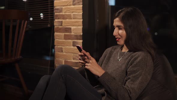 Brunette Is Sitting in a Bean Bag Chair in a Cafe in Evening Using Smartphone