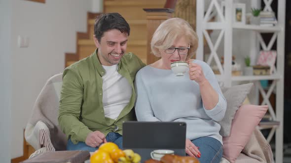 Positive Caucasian Adult Son and Senior Mother Sitting in Living Room Watching Movie Comedy Show