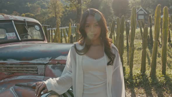 Young Asian Woman Stands By an Old Pickup Truck at Sunset and Looks at the Camera