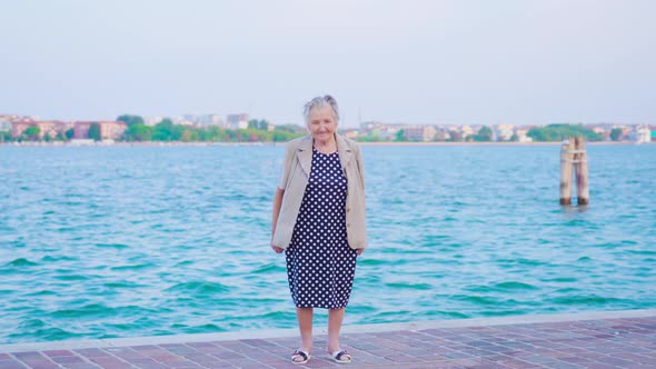 Old Woman Looks Into Camera Standing Against Venice Lagoon