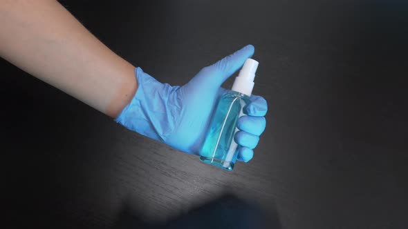 Hand in blue medical glove is holding hand sanitizer spray against black background. 