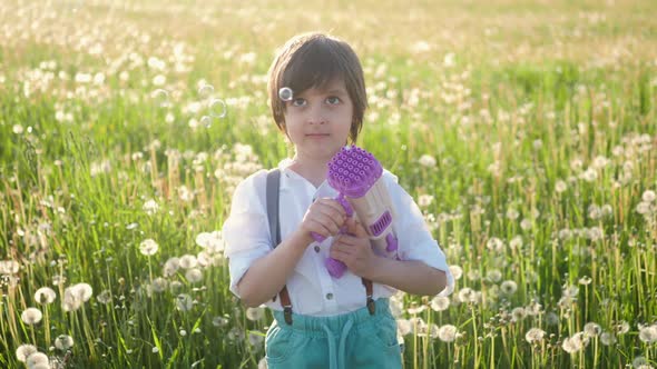 Portrait of a Five Year Old Boy in a Hat Stands on a Field of Dandelions and Shoots Soap Bubbles