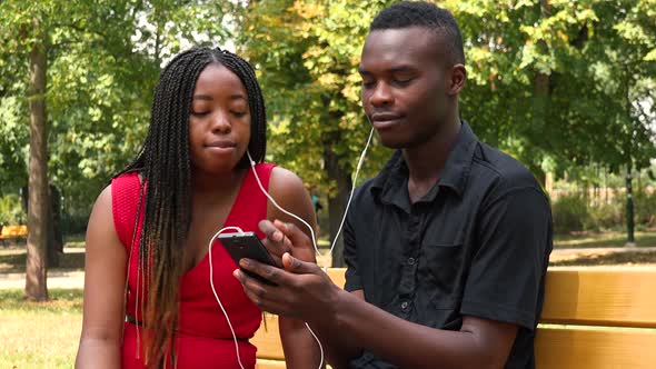 A Black Man and a Black Woman Sit on a Bench in a Park and Listen To Music on a Smartphone