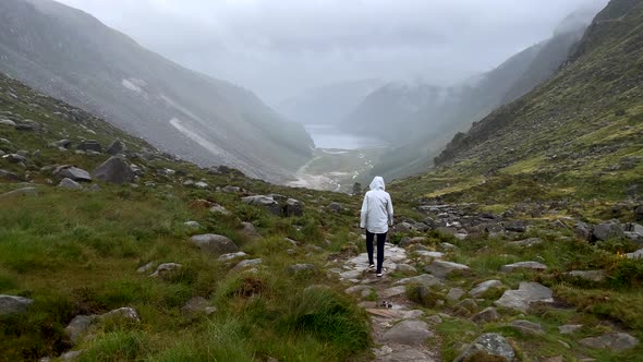 Rear view of female person with raincoat hiking on rocky path between giant mountains of Ireland dur