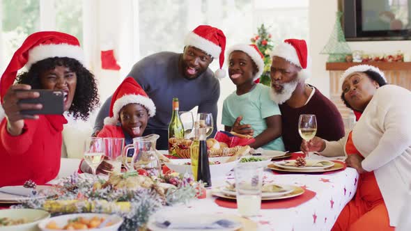 African american family in santa hats taking a selfie on smartphone while sitting on dining table ha