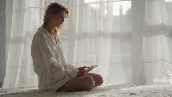 Portrait of Positive Caucasian Girl Sitting on Bed with Tablet and Smiling. Young Blond Woman