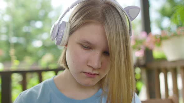 Teenage Girl Sits At Wooden Table In a Summer Cafe In Headphones With Phone