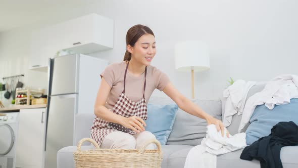 Asian service woman worker cleaning take messy dirty clothes into basket in living room at home.