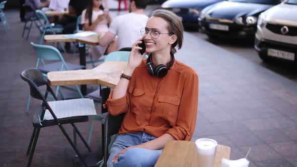 Happy Confident Blonde Woman in Denim and Orange Shirt Sitting on a Chair in a Street Cafe