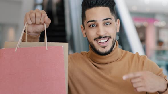 Happy Young Arab Man Buyer Standing in Mall Smiling Holding Shopping Bags in Hand Pointing