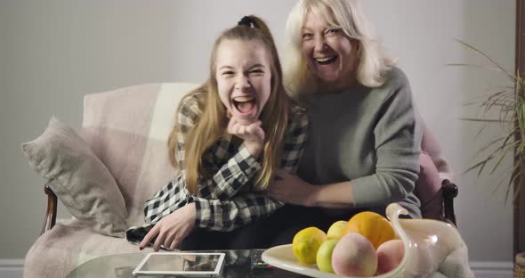 Portrait of Cheerful Caucasian Grandmother and Granddaughter Watching TV Together and Laughing