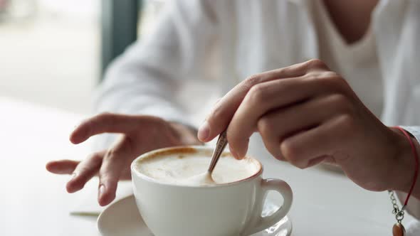A Hand Stirs A Cup Of Coffee With A Spoon, Cappuccino Coffee In A White Cup Stirring The Coffee Foam
