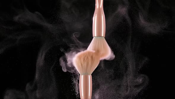 Two Makeup Brushes Collide and Cause a Swirl of Cosmetic Powder Particles Against a Black Background