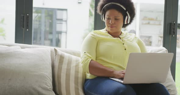 Video of happy plus size african american woman with headphones sitting on sofa with laptop