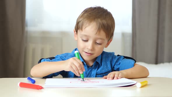 A Cute Boy Sits at a Table in a Room and Spends Time Painting with Colored Felt Pen.