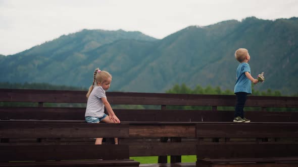 Boy with Wild Flowers Looks at Mountains Girl Sits on Bench