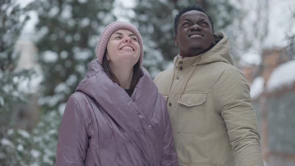 Joyful Happy Interracial Couple Standing in Snowfall Hugging and Smiling Looking at Camera