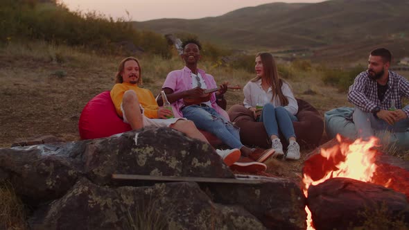 Group of International Friends are Sitting Around Campfire and One of Them is Playing Ukulele