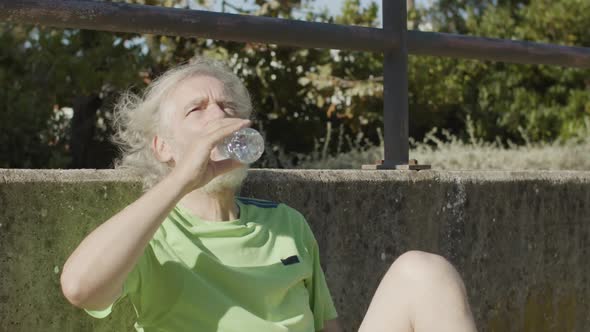 Tired Senior Man Sitting on Football Field and Drinking Water