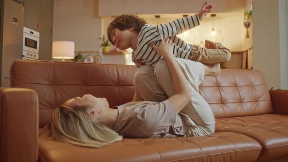 Happy Healthy Mother Holding Lifting Cute Little Preschool Child Son Up Playing Plane on Sofa at