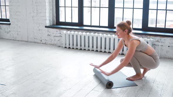 Cheerful young woman in stylish beige tracksuit unrolls rubber mat before training