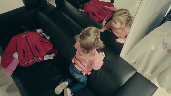 Little Child Girl in a Clothing Store Sits on the Couch in Front of a Mirror