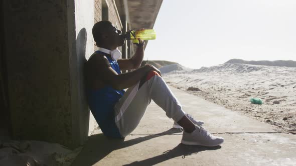 African american man sitting and drinking from water bottle taking break in exercise outdoors