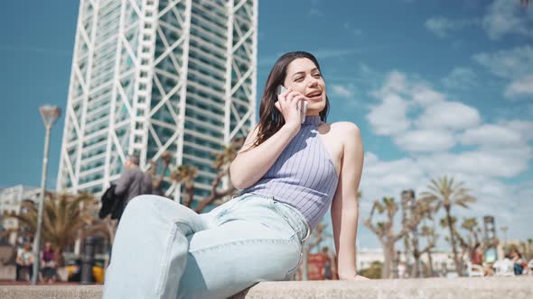 Cheerful brunette woman talking by mobile while sitting on the bench