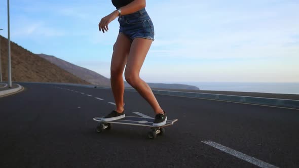 Girl in Shorts and Sneakers Skateboarding Along the Road on the Background of the Ocean and
