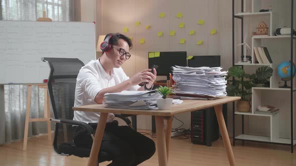 Asian Man With Headphones Listening To Music On Smartphone After Working With Documents