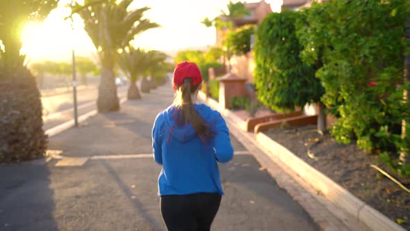 Woman Runs Down the Street Among the Palm Trees at Sunset Back View