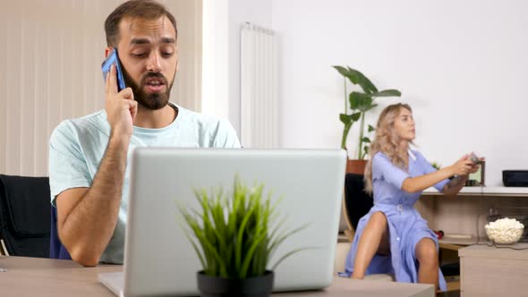 Man Talks on the Phone with a Laptop in Front While Her Wife Is Playing Video Games on a Console